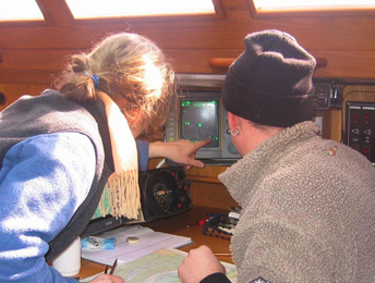 Man schaut einer blonden Frau und einer weiteren Person über die Schulter auf einen Radarschirm. Das Radarbild hat einen grau-schwarzen Hintergrund auf dem die Radarechos grün dargestellt sind. Auf dem Kartentisch davor liegt eine Seekarte.