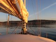 Blick über das Deck einer Segelyacht mit dem Mast im Mittelgrund in Ansteuerung auf Thorshavn, der Hauptstadt der Faröerinseln. Die Sonne steht tief und taucht alles in ein goldenes Licht.