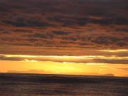 Unter einem von tiefhängenden Wolken bedeckten Himmel werden am Horizont befindliche Inseln in ein schemenhaftes goldenes Sonnenlicht getaucht, dass durch eine Wolkenlücke herabscheint. Die See ist spiegelglatt und nur von der Restdünung geschwungen.