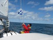 An Deck einer Segelyacht mitten auf dem Meer hockt ein Mann in roter Segeljacke an der Reling und zieht die schottische Gastlandflagge herab. Das Boot segelt bei Sonnenschein dahin.