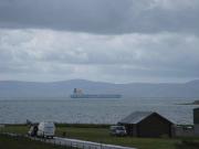 Unter wolkenverhangenem Himmel ankert ein blauer Tanker in Scapa Flow, einer großen Ankerbucht auf den Orkneyinseln. Im Hintergrund sieht man eine Hügelkette, im Vordergrund parken zwei Lieferwagen auf an saftig grünen Wiesen.