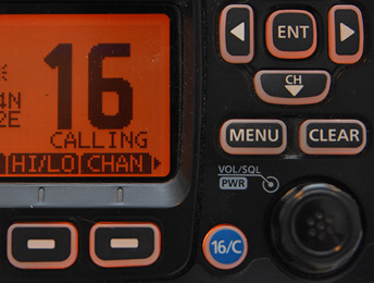 Ausschnitt eines orange hinerlegten Displays eines UKW-Funkgerätes. Auf dem Display ist groß die Zahl 16 für den allgemeinen UKW Not- und Anrufkanal 16 zu erkennen. Neben dem Display sind ein Drehknopf und einige Softkeys zur Bedienung des Funkgerätes.