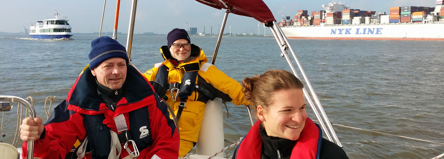 Drei Segelschüler am Heck unserer Segelyacht lächeln in Ölzeug bei sonnigem Wetter auf der Elbe in die Kamera. Im Hintergrund fährt ein großes Containerschiff vorbei und ein Ausflugsschiff überholt die Segelyacht.
