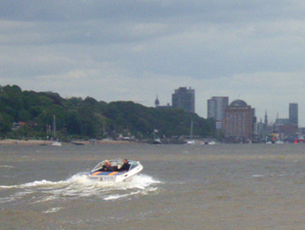 Ein offenes Motorboot mit zwei Personen fährt vor Övelgönne auf der brau-grau erscheinenden Elbe Richtung Hamburg. Im Hintergrund erkennt man schon die Hochhäuser und Türme der Innnenstadt
