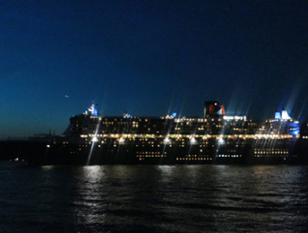 Die Silhouette eines hell erleuchteten Kruezfahrtschiffes auf der Elbe. Die zahlreichen Kabinenfenster sind erleuchtet, der Schornstein mit dem Logo der Reederei ist angestrahlt und die Decksbeleuchtung strahlt in die Nacht hinein.