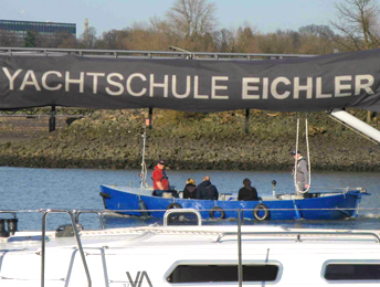 Unter dem Schriftzug Yachtschule Eichler ist unser Elektromotorboot E-Express auf dem Köhlfleet gegenüber der Lotsenstation Finkenwerder bei der Sportbootausbildung zu sehen. Im Vordergrund ist das Deck unserer Segelyacht zu sehen.