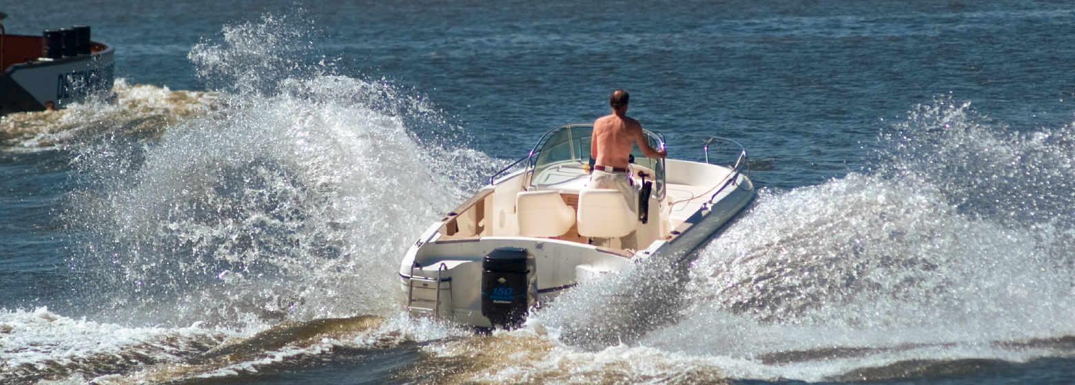 Ein offenes weißes Motorboot fährt mit hoher Geschwindigkeit durch das Wasser vom Betrachter weg, so dass die Gischt nach beiden Seiten weit wegspritzt. Am Ruder sieht man einen Mann von hinten mit nacktem Oberkörper in der strahlenden Sonne.