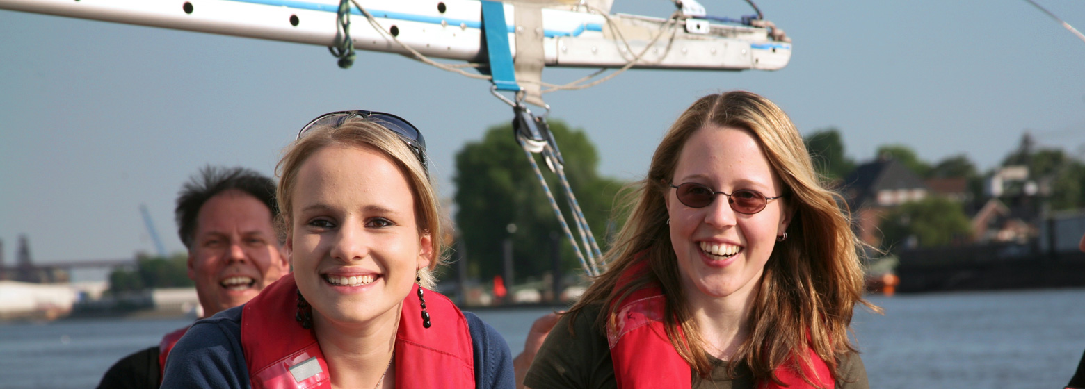 Im Vordergrund lächeln zwei junge Frauen in die Kamera, im Hintergrund lacht Robert Eichler. Sie genießen im Cockpit des Bootes das Segeln auf der Elbe.