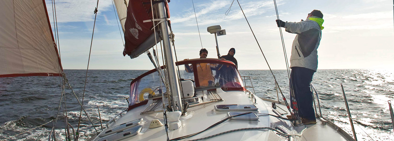 Eine moderne Segelyacht auf dem Meer von vorne nach achtern fotografiert. Die Sonne lässt das Wasser glitzern. Am Want in der Bootsmitte steht ein Mensch und schaut in Mast hinauf. Im Cockpit hinter der Sprayhood gucken zwei Leute nach vorne.